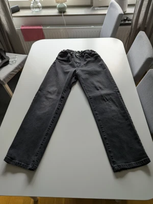 Svarta raka jeans med resårmidja - Säljer ett par svarta jeans med rak passform och resår i midjan. Klassisk femficksmodell med knapp och dragkedja framtill. Jeansen har en enkel och clean look som funkar till det mesta. Materialet är jeans och färgen är mörk svart.