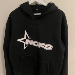 Svart hoodie med NOFS-tryck - Svart hoodie från NOFS med stor vit stjärna och logga på bröstet. Tröjan har huva, magficka och lång ärm. Perfekt för dig som gillar streetwear och vill ha en enkel men snygg look. Materialet är mjukt och känns skönt mot huden.
