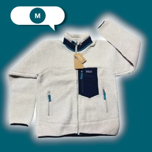 Vit teddyjacka från Patagonia M - Vit teddyjacka från Patagonia med marinblå detaljer och dragkedjor. Jackan har hög krage, tre fickor med dragkedja och ett coolt bröstfack i kontrastfärg. Materialet är mjuk fleece som håller dig varm och bekväm. Perfekt för kalla dagar. Storleken är M i herr men funkar utmärkt som unisex.