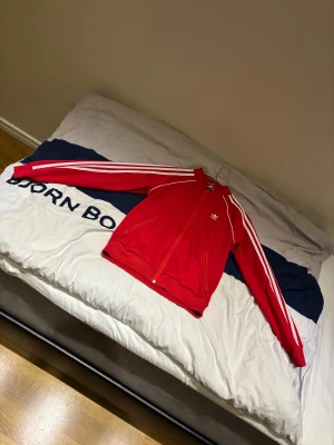 Röd Adidas vindjacka med vita ränder, - Säljer en helt oanvänd röd Adidas vindjacka med klassiska vita ränder längs ärmarna och Adidas-logga på bröstet. Jackan har dragkedja framtill, två fickor med dragkedja och ribbade muddar. Perfekt för dig som gillar sportig och retro stil.