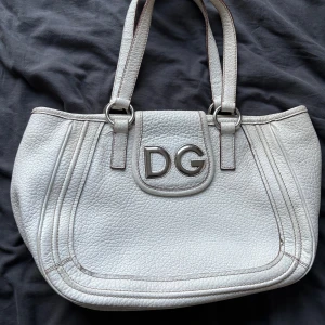 Äkta Dolce & Gabbana handväska i skinn - Snygg vit handväska från Dolce & Gabbana i äkta skinn med stora DG-metalldetaljer framtill. Väskan har dubbla handtag och coolt leopardmönstrat innerfoder. Perfekt för dig som vill sticka ut med en klassisk men edgy accessoar. Äkta och originellt köpt i Milano, köptes i somras på Vestiaire 