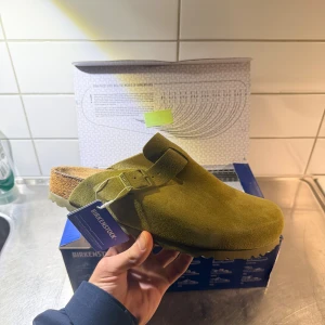 Olivgröna Birkenstock Boston sandaler - Säljer ett par olivgröna Birkenstock Boston sandaler i mocka med justerbart spänne och präglad logga på sidan. Skorna har en rund tå, platt sula och korkfotbädd för extra komfort. Perfekt för chill dagar och enkel att slänga på sig.