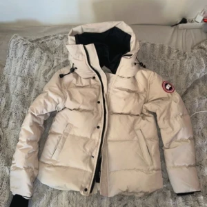 Beige Canada Goose pufferjacka - Säljer en beige pufferjacka från Canada Goose med ikonisk patch på ärmen. Jackan har huva, svarta detaljer, dragkedja och knappar framtill samt två sidofickor. Insidan är svart och jackan är dunfylld för extra värme. Perfekt för kalla dagar.