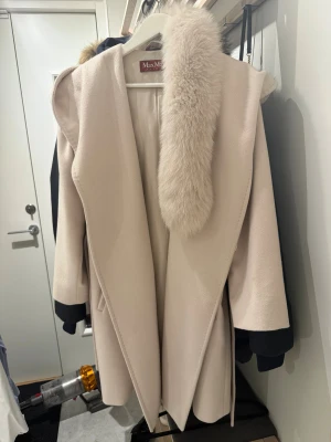 Beige Max Mara kappa med päls - Säljer en lyxig beige kappa från Max Mara med stor fluffig pälsdetalj vid kragen. Kappan har omlott-design, fickor i sidan och är tillverkad i mjuk ull. Perfekt för dig som vill ha en elegant och tidlös look under höst och vinter.