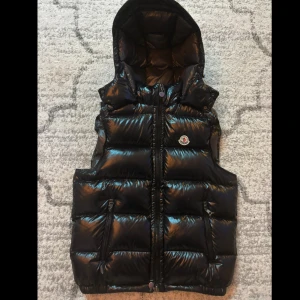 Moncler väst avtagbar luva - Svart, glansig dunväst från Moncler med avtagbar huva och ikonisk logga på bröstet. Västen har quiltad design, dragkedja framtill och praktiska fickor med dragkedja. Insidan har coolt Moncler-tryck och etiketter. Perfekt för att lyfta din streetstyle.
