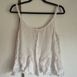 Vit volanglinne med knappar - Säljer ett vitt linne med smala axelband och volangkant nedtill. Toppen har knappar framtill och är tillverkad i ett lätt bomullstyg, perfekt för varma dagar. Enkel och söt design som passar till jeans eller kjol.
