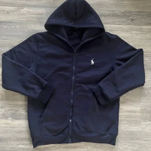 Ralph Lauren mörkblå hoodie - Mörkblå hoodie med dragkedja och vit logotyp på bröstet. Hoodien har fickor fram.   Den är inte använd bara att den saknar etikett och snören så annars nyskick! (Kontakta vid mer frågor eller bilder)