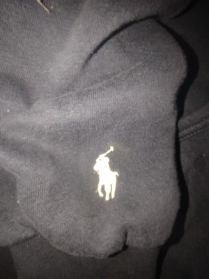 Marinblå hoodie från Polo Ralph Lauren - Snygg marinblå hoodie från Polo Ralph Lauren med stor vit logotyp på bröstet och klassisk polospelare broderad på ärmen. Tröjan har huva med snörning och en känguruficka framtill. I storlek Medium för herr
