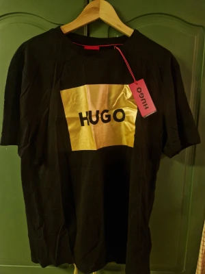 Svart t-shirt med guldtryck från Hugo - Oanvänd svart t-shirt från Hugo med ett stort guldigt tryck och svart HUGO-logga på bröstet. Det enda som behövs är en strykning. 