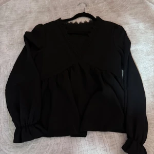Svart blus med spets och v-ringning - Snygg svart blus med v-ringning och spetsdetaljer runt halsen. Blusen har långa ärmar med resår vid ärmslut och en lös passform som ger en avslappnad look. Perfekt att styla med jeans eller kjol.