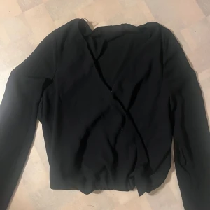 Svart omlottopp från BikBok XS - Säljer en svart omlottopp från BikBok i storlek XS. Toppen har lång ärm och en snygg v-ringning framtill. Enkel och stilren design som passar perfekt till jeans eller kjol. Materialet känns mjukt och följsamt.