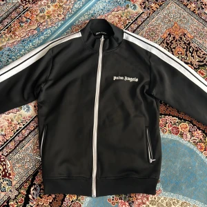 Svart Palm Angels track jacket L - Svart track jacket från Palm Angels med vita ränder längs ärmarna och dragkedja framtill. Jackan har ståkrage, två fickor med dragkedja och logga på bröstet. Tillverkad i bomull och polyester för en sportig vibe.