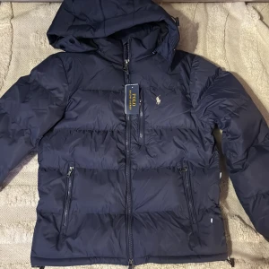 Mörkblå pufferjacka från Polo Ralph Lauren - Snygg mörkblå pufferjacka från Polo Ralph Lauren med huva och dragkedja framtill. Jackan har två sidofickor med dragkedja och klassisk Polo-logga på bröstet. Perfekt för kalla dagar och har en stilren, quiltad design.