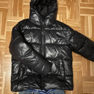 Svart pufferjacka  - Säljer en svart pufferjacka med glansig finish och huva. Jackan har dragkedja framtill och två sidofickor med dragkedja. Den är vadderad och har en loose passform, perfekt för kalla dagar. Enkel och stilren design som funkar till jeans och sneakers.