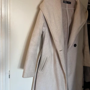 Beige kappa med luva, från Zara - Snygg beige kappa från Zara med bred krage och huva. Kappan har ett bälte i midjan, två fickor fram och stängs med knappar. Materialet är mjukt och känns lyxigt, perfekt för höst och vinter. Passformen är normal och ärmarna är långa. Har haft den i mindre än 1 år och den är knappt använd, den är lite nopprig då det har regnat på den men annars e den fortfarande jätte fin och nästan som ny