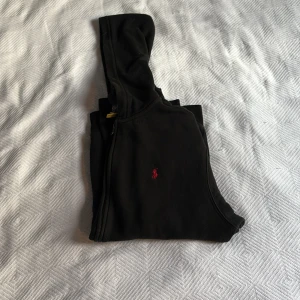 Svart hoodie från Polo Ralph Lauren - Svart hoodie från Polo Ralph Lauren med röd broderad logga på bröstet. Tröjan har huva, dragkedja framtill och två stora fickor. Tillverkad i mjuk bomull, perfekt för chill dagar. Klassisk och enkel design med långärmade ärmar. Folk som har S skulle möjligtvis kunna bära plagget också. Pris kan diskuteras!