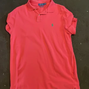 Röd pikétröja Polo Ralph Lauren - Snygg röd pikétröja från Polo Ralph Lauren i modellen Custom Slim Fit. Tröjan har klassisk krage, korta ärmar och två knappar vid halsen. Den ikoniska gröna Polo-loggan är broderad på bröstet. Perfekt för dig som gillar stilren och sportig look.