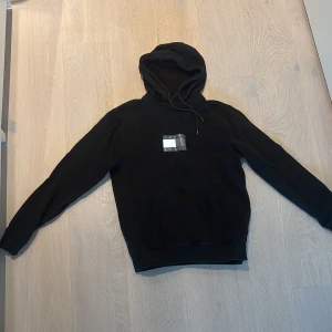 Svart hoodie Tommy Jeans M - Svart hoodie från Tommy Jeans i storlek M med klassisk logga på bröstet och liten detalj på ärmen. Perfekt för dig som gillar streetwear och enkel stil.