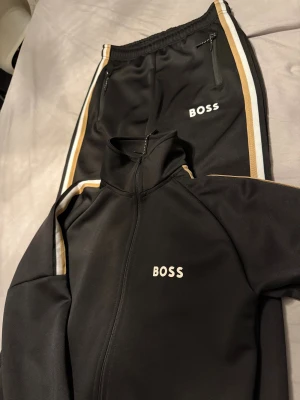 Svarta BOSS mjukisbyxor med revärer - Svarta mjukisbyxor från BOSS med vita och beige revärer längs sidorna. Byxorna har resår i midjan, dragkedjefickor och BOSS-logga på låret. Snygg sportig look, perfekta för chill eller träning.priset kan diskutera