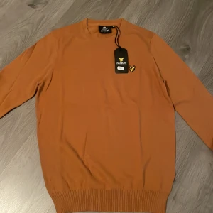Orange stickad tröja från Lyle & Scott - Snygg orange stickad tröja från Lyle & Scott med klassisk rund hals och ribbade muddar. Tröjan har lång ärm och den ikoniska gula örn-loggan broderad på bröstet. Perfekt för dig som gillar stilrena och tidlösa plagg.