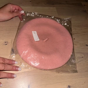 Rosa basker i ull mössa hatt för barn  - Säljer en superfin rosa basker i klassisk modell. Den är tillverkad i mjuk ull och har den ikoniska lilla knoppen på toppen. Perfekt accessoar för att lyfta din outfit med en fransk touch.En söt, rosa basker som ligger förpackad i plast. Perfekt för att addera en färgklick till din outfit.