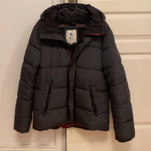 Svart pufferjacka från Garcia - Snygg svart pufferjacka från Garcia med huva och dragkedja framtill. Jackan har quiltad design, två fickor med dragkedja och en diskret logga på ärmen. Perfekt för kalla dagar och har en modern, boxig passform. Storlek 176 (motsvarar ungefär Medium) 