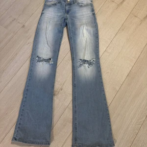 Ljusblå bootcut jeans med slitningar - Säljer ett par ljusblå jeans med bootcut passform och slitna detaljer över knäna. Jeansen har klassiska fickor och en snygg tvättad look.från Nelly storlek 36