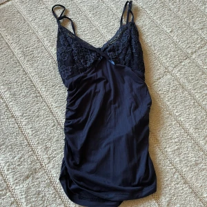 Mörkblå spetsdetaljerad linnetopp - Säljer en mörkblå linnetopp med smala axelband och v-ringad spets upptill med blommönster. Toppen har rynkade detaljer längs sidorna och en liten rosett framtill. Perfekt för dig som gillar feminina och stilrena plagg.