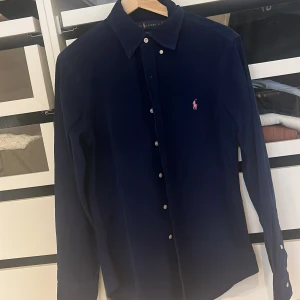 Marinblå manchesterskjorta Ralph Lauren - Slim fit marinblå skjorta från Ralph Lauren i mjuk manchester. Klassisk krage, vita knappar och långärmad design. Ikonisk rosa Polo-logga broderad på bröstet ger en snygg kontrast. Perfekt för dig som gillar stilrena och tidlösa plagg.