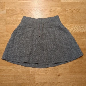 Grå stickad kortkjol från Divided - Grå stickad kortkjol från Divided med snyggt kabelmönster och bred ribbad midja med snörning. (Pris kan diskuteras)