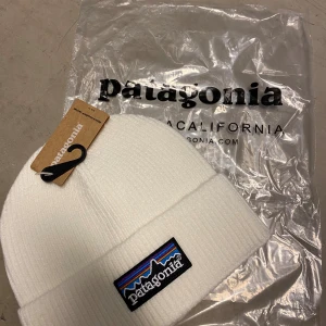 Vit ribbad mössa från Patagonia - Vit ribbad mössa från Patagonia med klassisk broderad logotyp i svart, blått, lila och orange framtill. Mössan har uppvikt kant och är tillverkad i mjukt stickat material som håller dig varm. Perfekt för kalla dagar och snygg till streetwear.