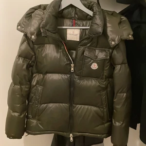 Mörkgrön pufferjacka från Moncler - Säljer en mörkgrön pufferjacka från Moncler med huva och klassisk logga på bröstet. Jackan har en quiltad design, dragkedja framtill och praktisk bröstficka med knapp. Insidan har en unik serietidningsetikett. Perfekt för kalla dagar.