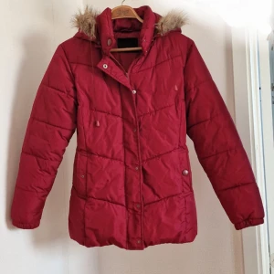 Röd quiltad pufferjacka med fuskpäls - Röd pufferjacka med quiltad design och huva med fuskpäls. Jackan har dragkedja och tryckknappar framtill, samt fickor med knappstängning. Perfekt för kalla dagar och har en snygg, klassisk look.