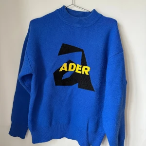 Blå stickad tröja från ADER Error - Säljer en blå stickad tröja från ADER Error med stor svart grafisk logga och gul text på bröstet. Tröjan har ribbad krage, muddar och lång ärm. Materialet är mjuk bomull och passformen är loose. Perfekt för dig som gillar statement-plagg och cool design. Size A1, equivalent to 36/38. Used but in good condition. 