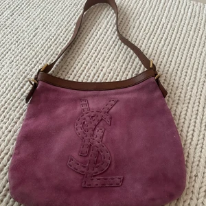 Yves Saint Laurent hobo Lila mocka  axelväska  - Säljer en snygg axelväska från Yves Saint Laurent i lila mocka med bruna skinn-detaljer och stort YSL-logo broderat på framsidan. Väskan har justerbar axelrem och innerfack med dragkedja. Perfekt för dig som vill ha en ikonisk och stilren väska.