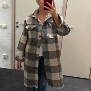 Rutig kappa - Snygg lång kappa i rutigt mönster från Cosy House. Jackan har stora bröstfickor med knappar, klassisk krage och knäppning framtill. Färgerna går i beige, grått och brunt. Skulle tro att storleken är S eller M