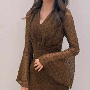 Brun långklänning med utsvängda ärmar - Lång brun klänning med transparenta, utsvängda ärmar och v-ringning. Klänningen har ett diskret prickigt strukturmönster i tyget och markerad midja. Materialet är lätt och ger en elegant känsla.