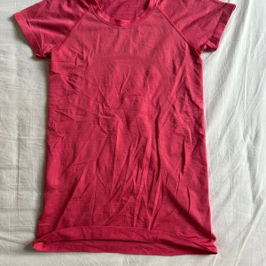 Rosa tränings-tshirt från Lululemon - Snygg rosa t-shirt från Lululemon med korta ärmar och rund hals. Perfekt för träning med sitt lätta och stretchiga material. T-shirten har diskreta ränder på ärmarna och en tight passform som sitter skönt på kroppen.Har klippt av taggen på insidan