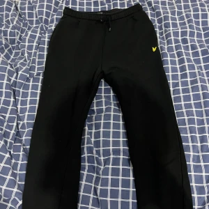 Svarta mjukisbyxor Lyle & Scott Junior - Svarta mjukisbyxor från Lyle & Scott Junior med elastisk midja och snörning. Byxorna har en gul broderad logga på vänster sida och en bakficka. Perfekta för chill dagar eller träning.