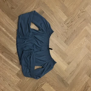Blå offshoulder topp från H&M Divided - Säljer en blå offshoulder topp från H&M Divided i storlek S. Toppen är croppad med långa ärmar och har smockdetaljer upptill och nedtill för en snygg passform. Perfekt för dig som gillar trendiga och bekväma plagg med lite extra edge.