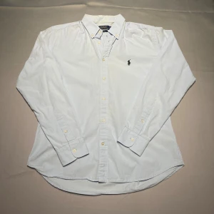 Ljusblå skjorta Polo Ralph Lauren Slim Fit - Klassisk ljusblå skjorta från Polo Ralph Lauren i slim fit-modell. Skjortan har button down-krage, långa ärmar och knappar framtill. Broderad svart logga på bröstet. En perfekt skjorta för en clean och stilren look. Skjortan har en liten missfärgning på insidan av kragen som knappt märks.