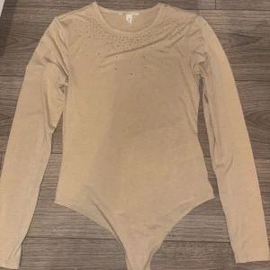 Beige långärmad topp med strass H&M - Snygg beige långärmad topp från H&M med rund halsringning och glittrande strassdetaljer över axlar och bröst. Mjuk och stretchig i bomullsmix, perfekt att styla med jeans eller kjol för en trendig look. Har aldrig används 