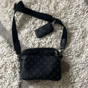 Louis Vuitton svart axelväska i skinn - Snygg svart axelväska från Louis Vuitton i skinn med klassiskt monogrammönster. Väskan har bred axelrem med logga, dragkedja och tillhörande liten plånbok. Perfekt för dig som vill ha en stilren och lyxig accessoar.