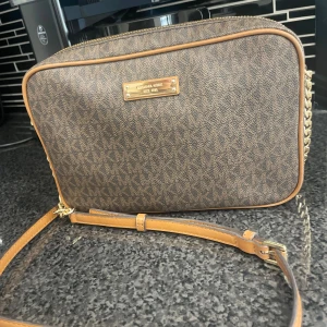 Brun axelväska Michael Kors - Snygg brun axelväska från Michael Kors med MK-logga över hela väskan. Väskan har guldfärgade kedjedetaljer på axelremmen och justerbar läderrem. Rektangulär form och dragkedja upptill. Perfekt storlek för det viktigaste.