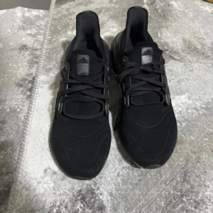 Svarta Adidas Ultraboost sneakers - Säljer ett par helsvarta Adidas Ultraboost sneakers med klassiska tre ränder på sidan och snörning. Ovandelen är i mesh och syntet, och sulan är i mjukt, stötdämpande material för extra komfort. Perfekta för dig som gillar stilrena och sportiga skor.