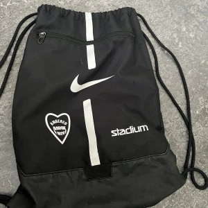 Svart gympåse från Nike x Stadium - Svart gympåse med dragsko från Nike och Stadium, Praktisk dragkedjeficka framtill. Perfekt för träning eller skola.