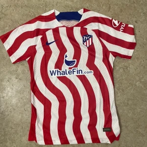 Atletico Madrid matchtröja Nike - Säljer en officiell Atletico Madrid fotbollströja från Nike med unikt böljande rödvitt randigt mönster. Tröjan har korta ärmar, klubbmärke på bröstet och sponsorer tryckta fram och bak. Materialet är lätt och ventilerande polyester, perfekt för träning eller match.
