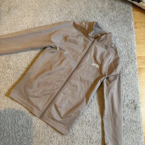 Beige träningsjacka från AYBL XS - Säljer en beige träningsjacka från AYBL i storlek XS. Jackan har hel dragkedja, hög krage och tumhål vid ärmsluten. Tillverkad i mjukt och stretchigt material som passar perfekt till träning. Enkel och stilren design med logga på bröstet.