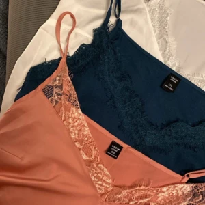 Spetslinnetopp från SHEIN i blått och aprikos - Två snygga linnetoppar från SHEIN, en i marinblått med spetskant och en i aprikos med spetsdetaljer. Båda har tunna axelband och ärmlös design, perfekta för att ge en chic vibe. Materialet är mjukt och lent, troligen polyester med spets. Säljes tillsammans!
