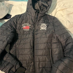 Svart pufferjacka Vinson Polo Club S - Svart pufferjacka från Vinson Polo Club med broderade loggor på bröstet och huva. Jackan har quiltad design, dragkedja framtill och ribbade muddar. Materialet är nylon på utsidan och polyesterfoder. Perfekt för kyliga dagar.